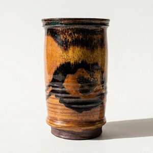 May include: A handmade ceramic tumbler with a cylindrical shape. The exterior features a blend of brown, black, and yellow hues, creating a marbled effect. The base and rim are a darker shade of brown. The tumbler is suitable for beverages.