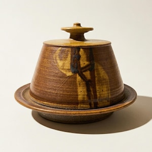 May include: A brown ceramic butter dish with a lid. The dish has a round base and a conical top with a small knob. The lid and dish are decorated with brown and yellow abstract designs.