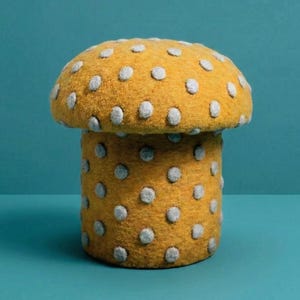 May include: A whimsical, mushroom-shaped decorative object. The mushroom has a mustard-yellow cap and stem, both adorned with evenly spaced, light gray polka dots. The object is set against a teal background, creating a playful and inviting aesthetic.