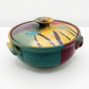 May include: A handmade ceramic lidded bowl with a colorful design. The bowl features a teal base, a maroon rim, and a lid with yellow, teal, and maroon accents. The lid is decorated with black lines and blue floral designs.