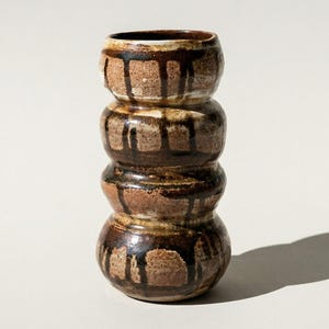 May include: A handcrafted ceramic vase with a stacked design. The vase features a dark brown and tan color scheme with dripped glaze details. The vase is approximately 6 inches tall.