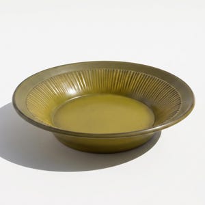 May include: A round, olive-green ceramic plate with a slightly raised rim. The rim features a textured, ridged design, while the center of the plate is smooth. The plate is set against a white background, casting a shadow.