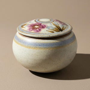 May include: A small, round ceramic jar with a lid. The jar is off-white with blue and yellow stripes around the middle. The lid is decorated with pink flowers and green leaves. The jar is likely used for storage.