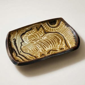 May include: A rectangular ceramic dish with a decorative floral design. The dish features a brown and tan color palette with black accents. The edges are dark brown, and the interior has a detailed pattern. The dish is likely used for serving or display.