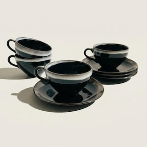 May include: A set of black ceramic teacups and saucers. The cups have a rounded shape with a handle and a light-colored rim. The saucers are round and match the cups. The set includes four cups and saucers.
