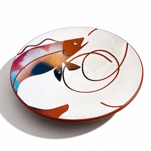 May include: A round ceramic plate with a white base featuring a stylized brown fish illustration. The fish design incorporates blended patches of blue, pink, and orange colors. Thin, dark brown curved lines swirl across the surface of the plate.