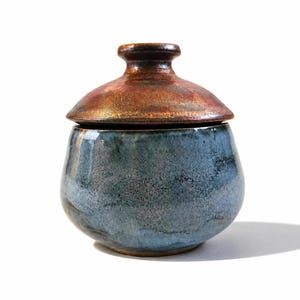 May include: A ceramic jar with a rounded body and a matching lid. The base of the jar features a mottled blue and grey glaze with a textured finish. The lid has a smooth, rounded knob and is finished in a metallic copper and bronze glaze.