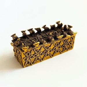 May include: A rectangular, gold-colored decorative box with intricate floral and geometric patterns. The box has a series of small, ornate figures along the top. The overall design suggests a vintage or antique aesthetic, suitable for storing small items.