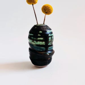 May include: A small, handcrafted ceramic vase with a dark, textured exterior and a splash of green and blue glaze. Two yellow, spherical flowers are placed inside the vase. The vase is a decorative item.