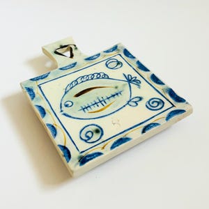 May include: Square ceramic cutting board with a handle, featuring a hand-painted fish design. The board has a white base with blue and gold accents, including a border of blue dots and swirls. The fish is depicted in blue and gold.