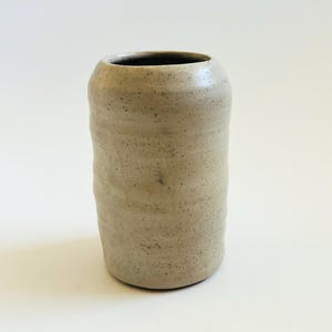 May include: A handmade, cylindrical ceramic vase with a textured, speckled surface. The vase is a light beige color with a slightly irregular shape, and a dark interior. The vase is suitable for displaying flowers or as a decorative item.