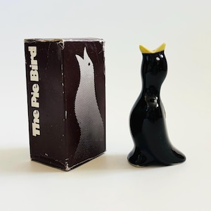 May include: A black ceramic bird figurine with a yellow beak, next to a brown box. The box has the text "The Pie Bird" and a white bird illustration. The figurine is glossy and has a simple, stylized design.