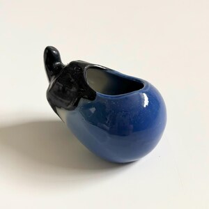 May include: A ceramic eggplant-shaped container with a glossy finish. The body is a deep blue, and the stem is black. The opening is a small oval. This decorative piece could be used as a small planter or a unique kitchen accent.