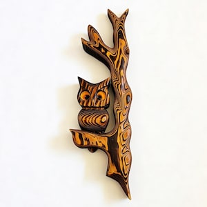 May include: A wooden owl sculpture perched on a branch. The owl has large, orange eyes and is carved with detailed wood grain patterns. The tree branch and trunk are also carved from wood, with a similar grain pattern and a dark brown color.