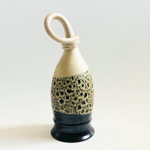May include: A ceramic bottle stopper with a loop handle. The top is off-white, transitioning to a textured, speckled brown and black midsection, and a dark blue base. The loop handle is off-white.