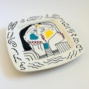 May include: A white, square ceramic plate with a colorful abstract face design. The face features black outlines, a black hair, blue, yellow, and red accents. The plate is decorated with black squiggles around the edges.