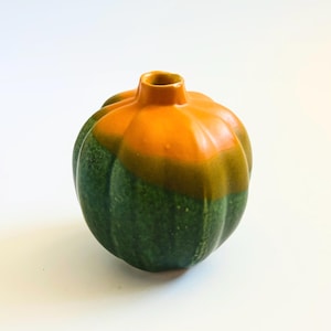 May include: A ceramic vase shaped like an acorn squash. The vase is green on the bottom, transitioning to a band of olive green, and topped with a bright orange color. The vase has a small opening at the top.
