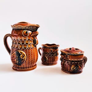 May include: A set of three brown ceramic kitchenware pieces. The largest is a pitcher with a rooster design, and the other two are a small mug and a lidded jar. The pieces have a glossy finish and are decorated with intricate patterns.