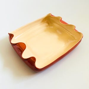 May include: A square ceramic serving dish with a wavy rim. The interior is a light beige color, transitioning to a golden hue in the center. The exterior and rim are a glossy, deep red.