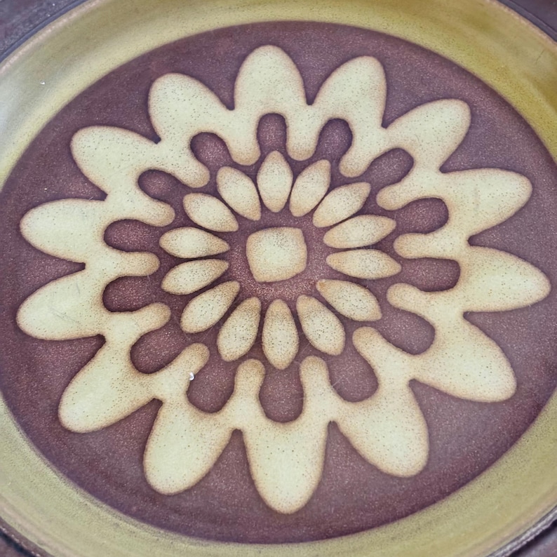 K&ouml;nnte beinhalten: Ein Keramikteller mit floralem Design. Der Teller hat ein zentrales Blumenmuster in Creme und Braun, umgeben von einem dunkelbraunen Hintergrund und einem hellgr&uuml;nen Rand. Das Design ist symmetrisch und detailliert.