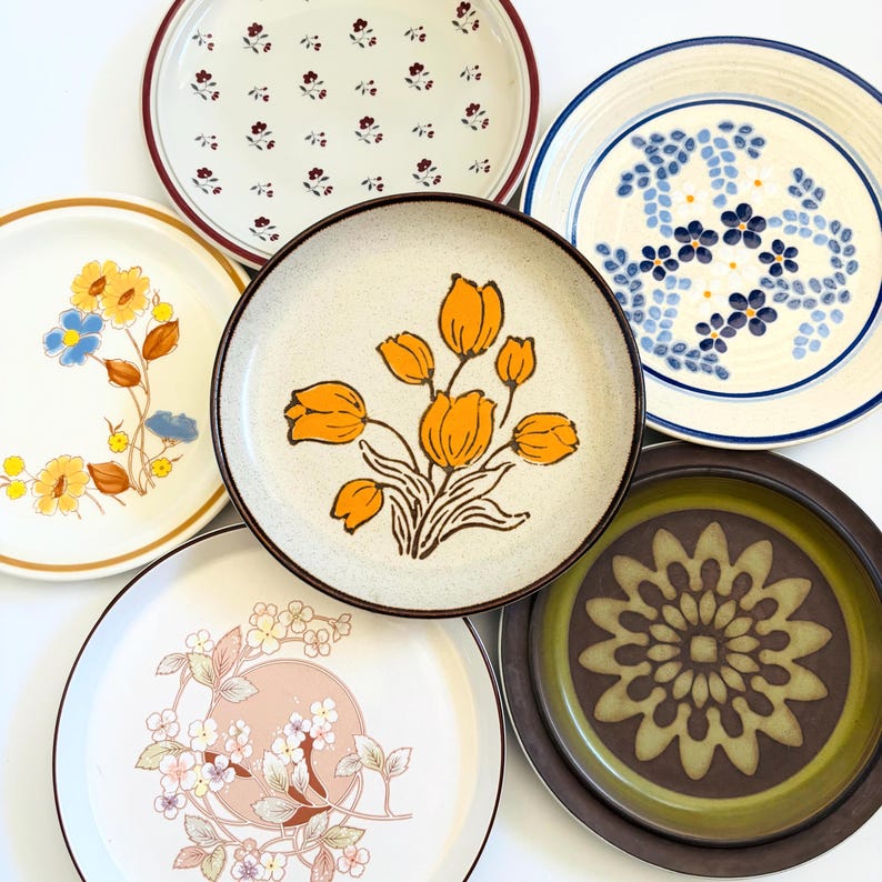 K&ouml;nnte beinhalten: Eine Sammlung von Vintage-Keramiktellern mit verschiedenen floralen Designs. Die Teller weisen eine Reihe von Farben auf, darunter Gelb, Blau, Braun und Wei&szlig;. Einige Teller haben einen durchgehenden Rand, andere aufwendige Muster.