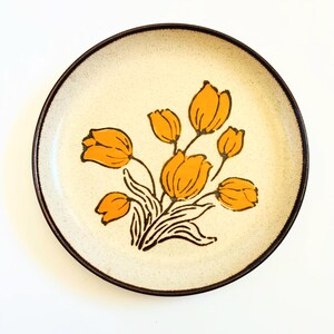 K&ouml;nnte beinhalten: Ein runder Keramikteller mit einer cremefarbenen, gesprenkelten Oberfl&auml;che und einem dunkelbraunen Rand. Der Teller zeigt ein Blumendesign aus orangen Tulpen mit schwarzen Stielen und Bl&auml;ttern. Ein dekorativer Teller im Vintage-Stil.