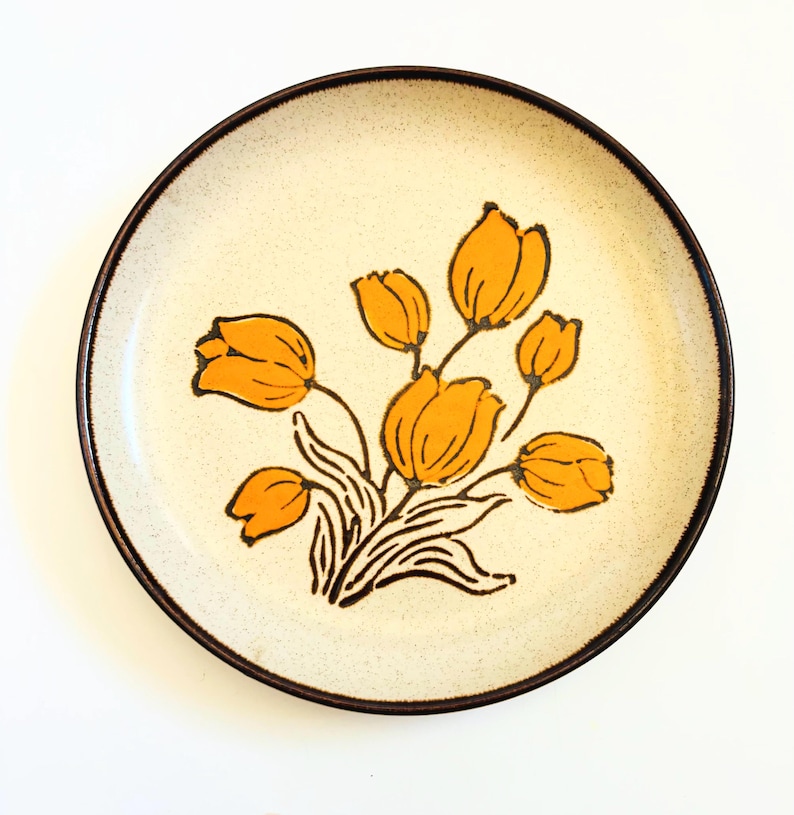 K&ouml;nnte beinhalten: Ein runder Keramikteller mit einer cremefarbenen, gesprenkelten Oberfl&auml;che und einem dunkelbraunen Rand. Der Teller zeigt ein Blumendesign aus orangen Tulpen mit schwarzen Stielen und Bl&auml;ttern. Ein dekorativer Teller im Vintage-Stil.
