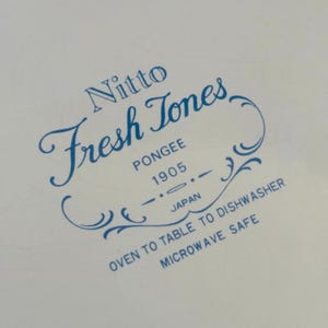 K&ouml;nnte beinhalten: Nahaufnahme einer wei&szlig;en Oberfl&auml;che mit blauem Text. Der Text lautet "Nitto Fresh Tones PONGEE 1905 JAPAN OVEN TO TABLE TO DISHWASHER MICROWAVE SAFE". Der Text ist in einer dekorativen Schriftart.