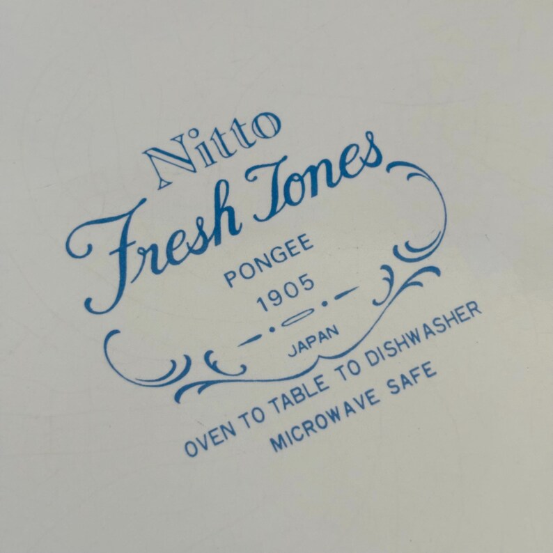 K&ouml;nnte beinhalten: Nahaufnahme einer wei&szlig;en Oberfl&auml;che mit blauem Text. Der Text lautet "Nitto Fresh Tones PONGEE 1905 JAPAN OVEN TO TABLE TO DISHWASHER MICROWAVE SAFE". Der Text ist in einer dekorativen Schriftart.