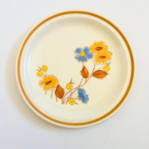 K&ouml;nnte beinhalten: Ein runder, cremefarbener Teller mit braunem Rand und floralem Design. Die Blumen sind gelb und blau mit braunen Stielen und Bl&auml;ttern. Der Teller ist im Vintage-Stil und eignet sich zum Servieren von Speisen.