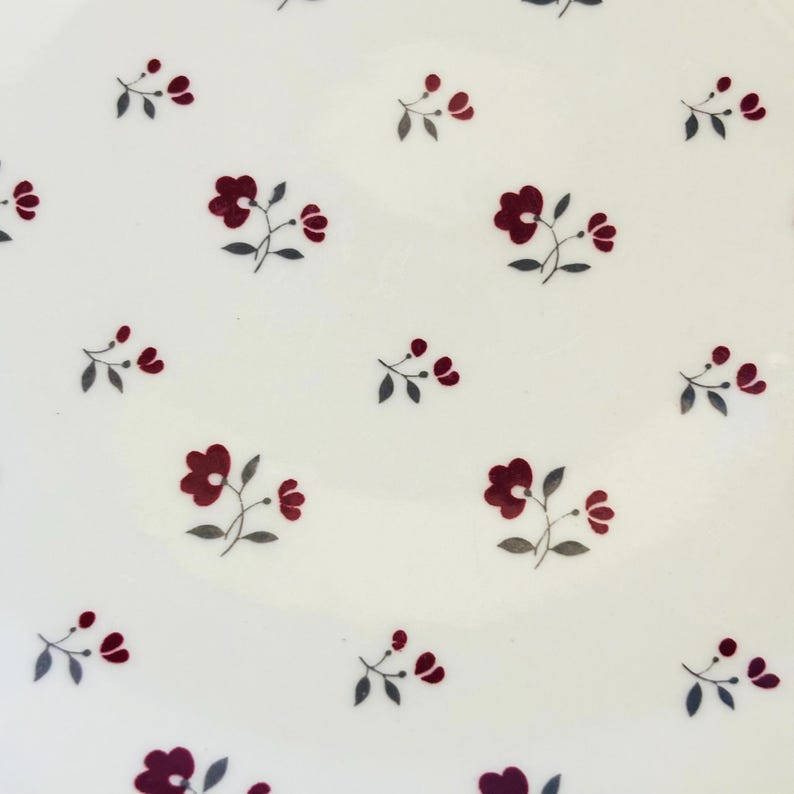 K&ouml;nnte beinhalten: Wei&szlig;e Keramikplatte mit einem sich wiederholenden Muster aus kleinen, dunkelroten Blumen und grauen Bl&auml;ttern. Die Blumen sind in Gruppen angeordnet und bilden ein zartes Design. Die Platte hat eine schlichte, saubere &Auml;sthetik.