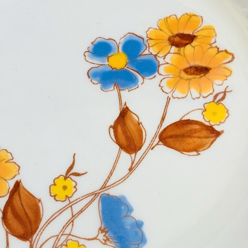 K&ouml;nnte beinhalten: Nahaufnahme eines wei&szlig;en Keramiktellers mit floralem Design. Die Blumen sind blau und gelb mit braunen Stielen und Bl&auml;ttern. Das Design ist im Vintage-Stil.
