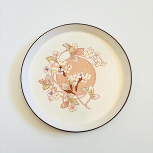 K&ouml;nnte beinhalten: Runder, cremefarbener Keramikteller mit braunem Rand. Der Teller hat ein Blumendesign mit einem pfirsichfarbenen Kreis in der Mitte, umgeben von zarten Blumen und Bl&auml;ttern in Rosa, Gelb und Braun.