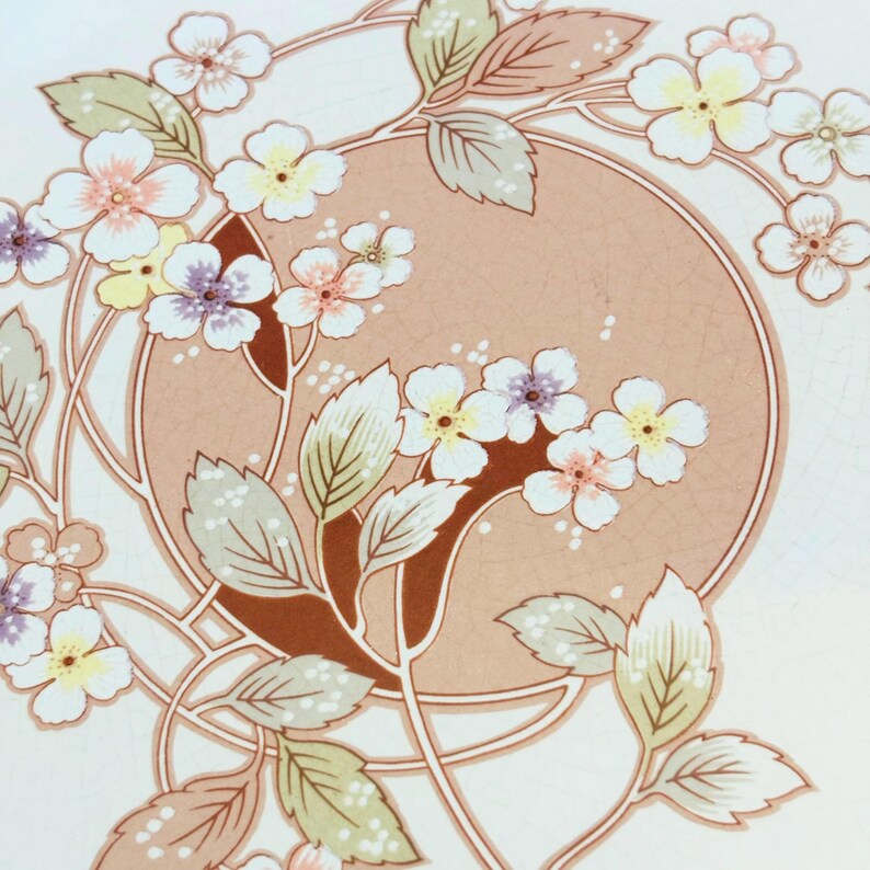 K&ouml;nnte beinhalten: Blumenmuster mit einem pfirsichfarbenen Kreis in der Mitte, umgeben von wei&szlig;en Bl&uuml;ten mit gelben, rosa und violetten Zentren. Das Design umfasst gr&uuml;ne und hellbraune Bl&auml;tter und Stiele, alles vor einem wei&szlig;en Hintergrund.