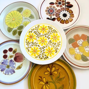May include: A collection of six vintage ceramic plates with floral designs. The plates feature various colors, including yellow, brown, blue, and green, with different floral patterns. The plates are arranged on a white surface.