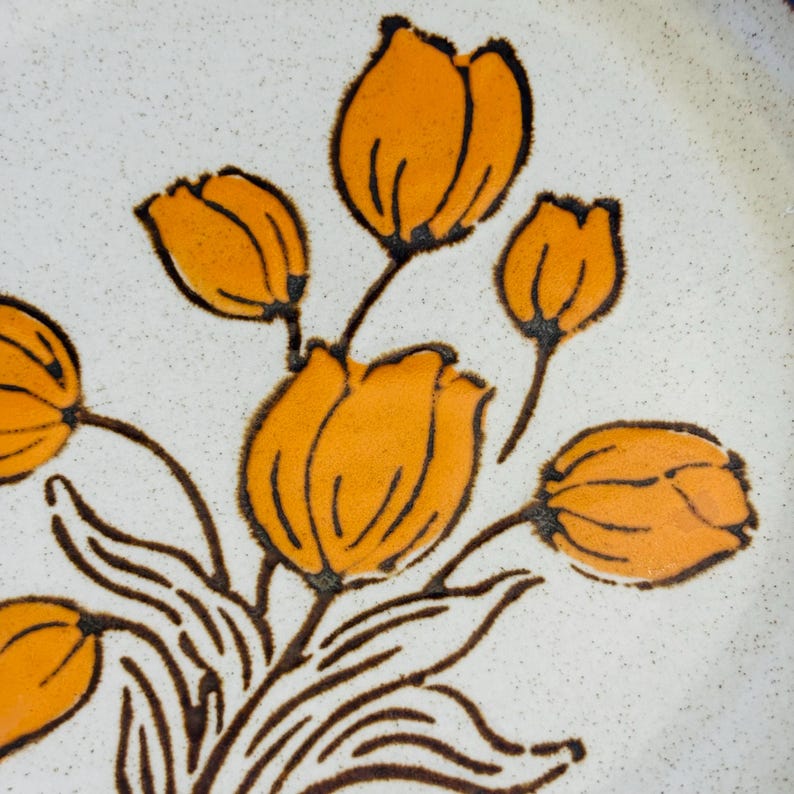 K&ouml;nnte beinhalten: Nahaufnahme eines Keramiktellers mit floralem Design. Der Teller zeigt orangefarbene, tulpenartige Blumen mit braunen Umrissen und Stielen auf einem gesprenkelten wei&szlig;en Hintergrund. Das Design wiederholt sich.