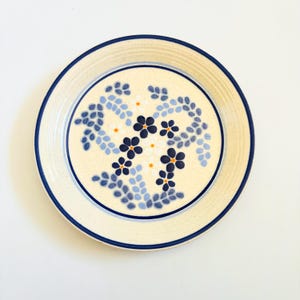 K&ouml;nnte beinhalten: Ein runder, cremefarbener Teller mit blauem Blumendesign. Der Teller hat einen dunkelblauen Rand und einen hellblauen Innenring. Das Blumenmuster zeigt dunkelblaue und hellblaue Blumen und Bl&auml;tter.