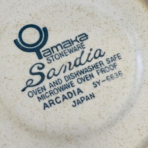 K&ouml;nnte beinhalten: Nahaufnahme eines Abschnitts eines Keramiktellers mit dem Text "Yamaka STONEWARE Sandia" in Dunkelblau. Der Teller ist cremefarben mit braunen Sprenkeln. Der Text weist auch darauf hin, dass er ofen- und sp&uuml;lmaschinenfest ist und in Japan hergestellt wurde.