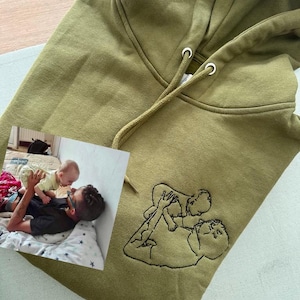 Könnte beinhalten: Olivgrüner Kapuzenpullover mit Kordelzugkapuze und Kängurutasche. Der Kapuzenpullover hat ein gesticktes Design einer Person, die ein Baby hält. Auf dem Kapuzenpullover befindet sich ein Foto einer Person und eines Babys.