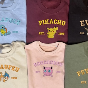 May include: Embroidered sweatshirts in various colors, each featuring a different Pokémon character and name. The designs include "CARAPUCE," "PIKACHU," "EVOLI," "DRACAUFEU," "RONDOUDOU," and "PSYKOK," with "EST. 1996" below each.