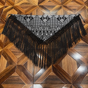 Puede incluir: Un chal triangular con un patrón geométrico en blanco y negro con detalles bordados intrincados. Los bordes están rematados con un fleco negro largo y grueso que cuelga de la parte inferior y los lados, creando una apariencia texturizada.
