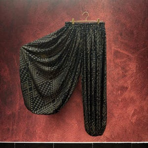 May include: Black harem trousers featuring a gold geometric pattern throughout the fabric. The trousers have a high, elasticated waistband and a voluminous, draped silhouette that gathers at the ankles, suspended from a gold-toned metal clothes hanger.