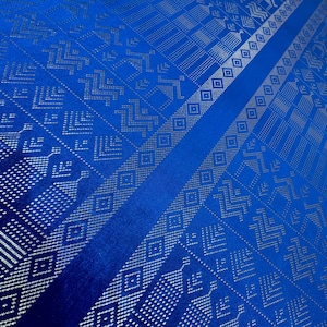 Puede incluir: Primer plano de una tela azul vibrante con intrincados patrones geométricos plateados. El diseño presenta rayas diagonales y formas repetidas, creando una apariencia texturizada. El brillo de la tela sugiere un material lujoso.