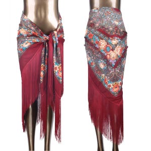 May include: A deep burgundy wrap skirt featuring a vibrant floral and paisley pattern in shades of teal, orange, and cream. The garment is finished with long, matching burgundy fringe along the hemline, creating a layered and textured appearance.