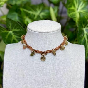 May include: A handmade necklace with a brown woven cord, decorated with green and tan gemstones. A small, round, gold-coloured pendant hangs from the centre. The necklace is displayed on a white mannequin head.