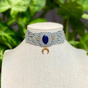 Peut inclure: Un collier ras du cou en macramé gris avec une grosse pierre ovale bleu foncé au centre. Le collier comporte des accents dorés et un pendentif en forme de croissant de lune doré. Le collier est présenté sur un mannequin.