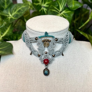 Puede incluir: Un collar gargantilla de macramé gris con detalles turquesa y cuentas color bronce. El collar presenta un colgante rojo y turquesa. El collar se exhibe en una cabeza de maniquí blanca.