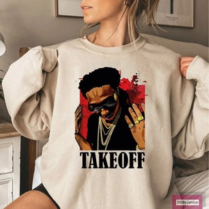Op de afbeelding: Beige sweatshirt met een afbeelding van een persoon met een zonnebril en gouden sieraden, tegen een rode achtergrond. Het woord "TAKEOFF" is in vette zwarte letters onder de afbeelding gedrukt. De sweatshirt heeft lange mouwen.