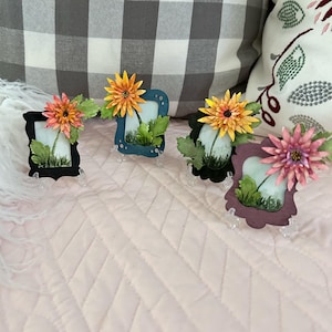 May include: Four miniature framed artworks, each featuring a floral arrangement with a colorful flower and green leaves. The frames are in black, teal, and purple, displayed on clear stands. The background includes a pink quilted surface and decorative pillows.