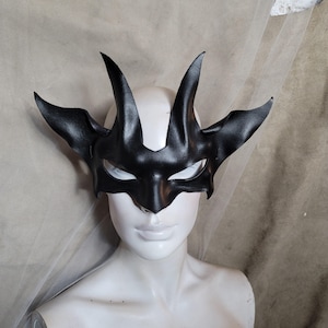 Könnte beinhalten: Eine schwarze Ledermaske mit einem gehörnten Design. Die Maske hat Augenausschnitte und wird auf einem weißen Schaufensterkopf präsentiert. Die Maske hat ein gotisches oder Fantasy-Thema.